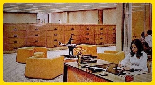 Old photo from the 70s showing an office decor in orange and tan with rows of wide, wooden filing cabinets. A woman in white with mid-length black hair sits at a desk close to camera, writing on a pad of paper. An indecipherable object sits on a small table surrounded by four orange, upholstered chairs.