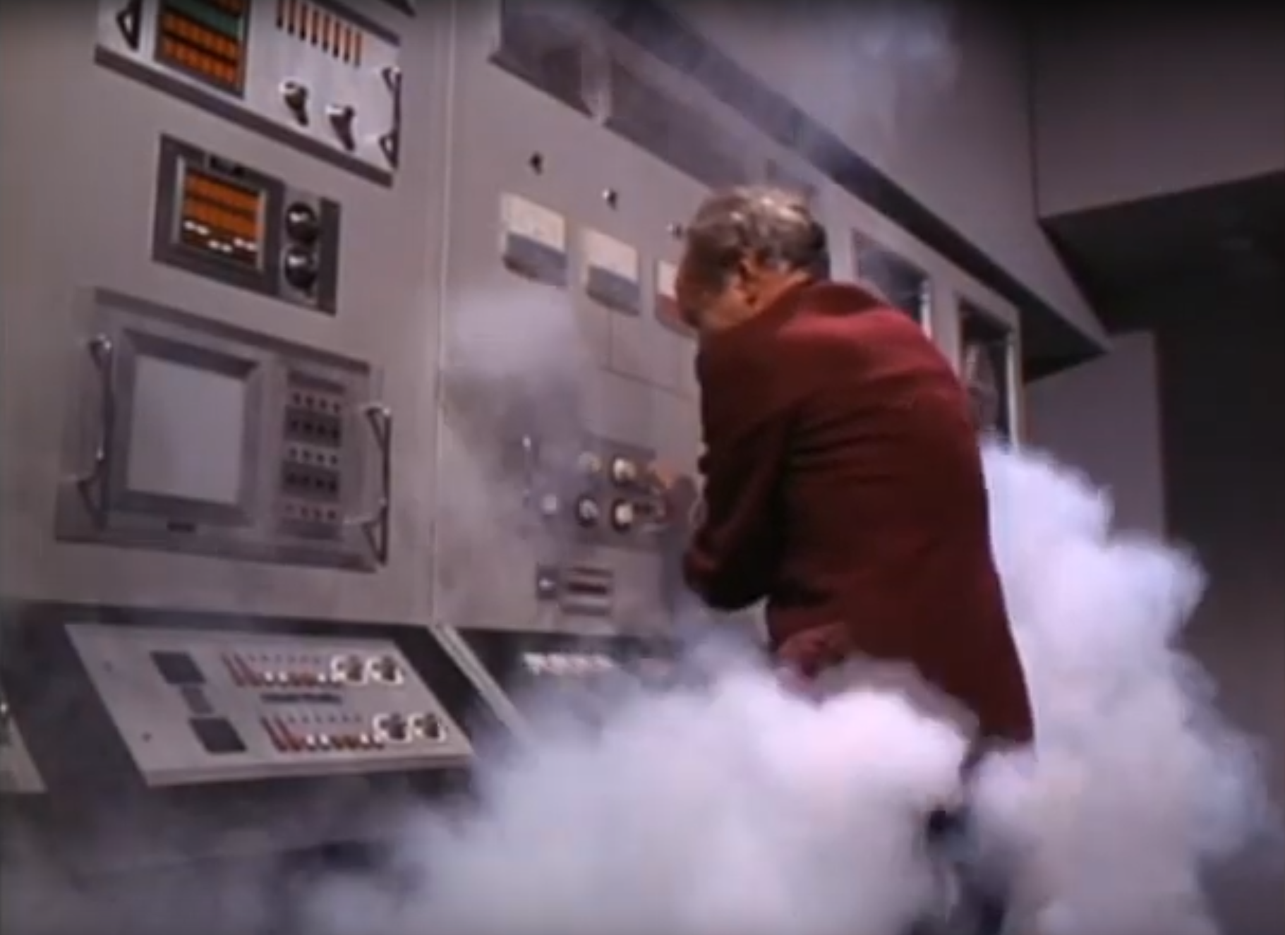 Screen capture from The Prisoner television show. A complex computer system has apparently burned itself out, smoke billowing from its cooling vents. A middle-aged man in a burgundy jacket desperately fights the controls to save the system.