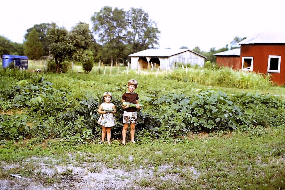 A young boy and his younger sister hold giant zucchini plucked from the very large vegetable garden behind them. They are smiling. In the background, two horse barns can be seen, as well as a horse trailer. The picture seems to have been shot in the morning, with a slightly overcast sky.