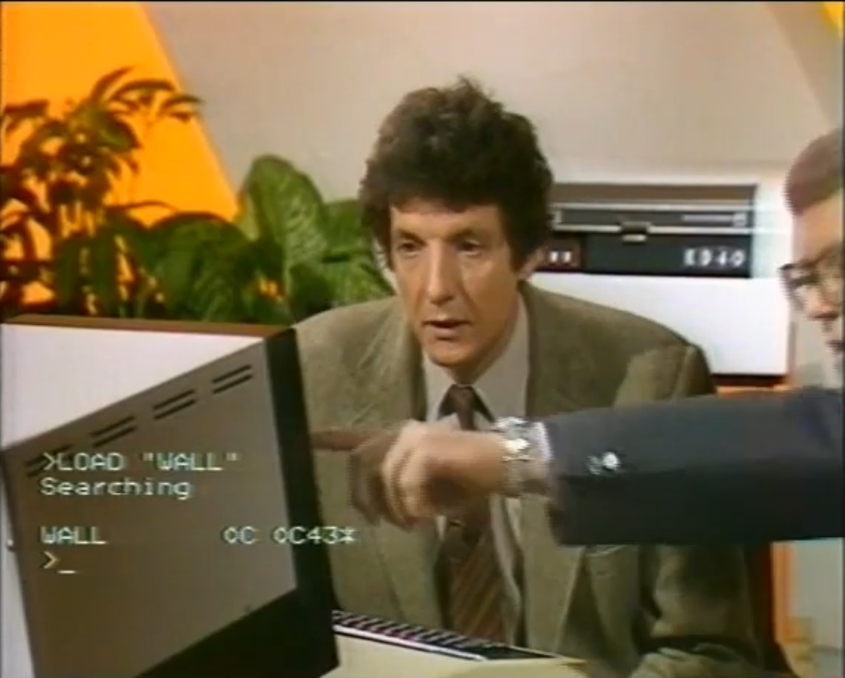 A still frame from the BBC’s The Computer Programme. Presenter Chris Serle sits at a computer looking confused while his co-host off-screen points at the monitor. Superimposed on the live image, text from the computer screen shows a BASIC prompt loading a program called ‘WALL.’