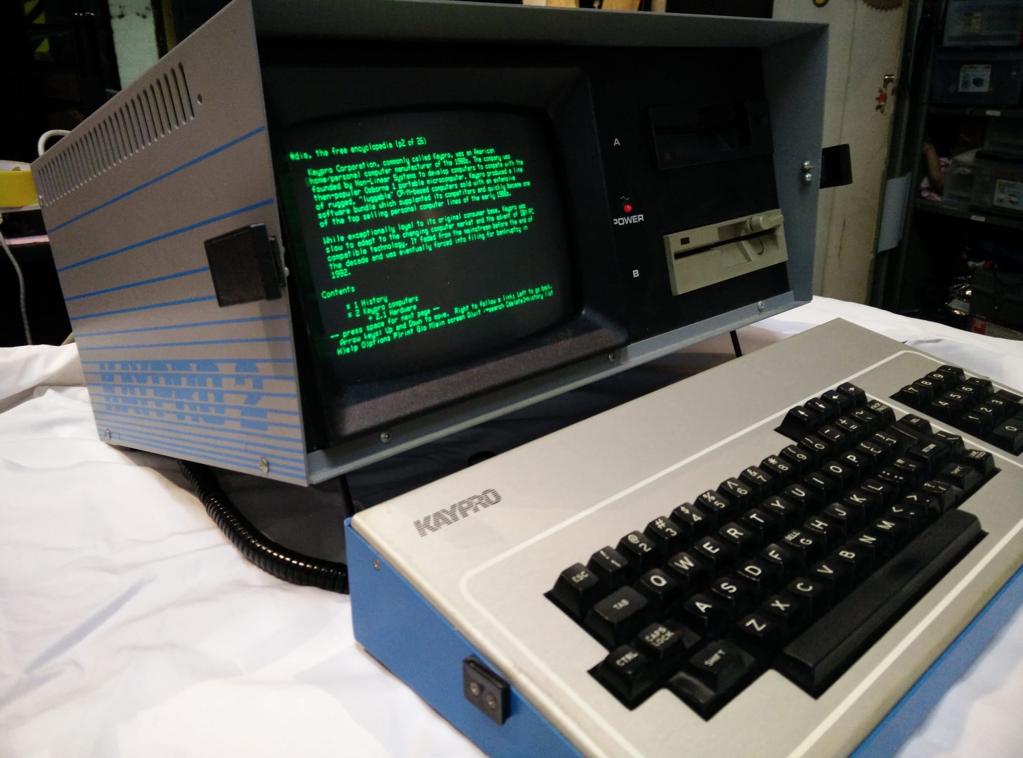 Kaypro II portable CP/M computer with its gunmetal grey metal case open, showing a built-in green monochrome CRT and dual 5.25-inch floppy drives. The detachable keyboard, branded “Kaypro,” sits in front, connected by a coiled cable. The screen displays text-only Wikipedia through some trick of modern programming.