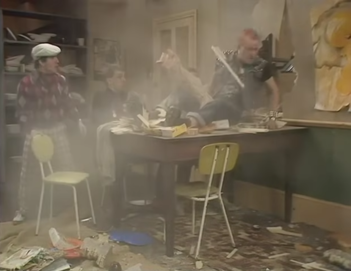 Screen capture from episode one of The Young Ones. Punk rocker Vyvyan has made his first entrance, crashing through a wall onto the kitchen table. Mike in golfing attire, Rik in all black, and Neil in drab hippie clothes look on in shock.